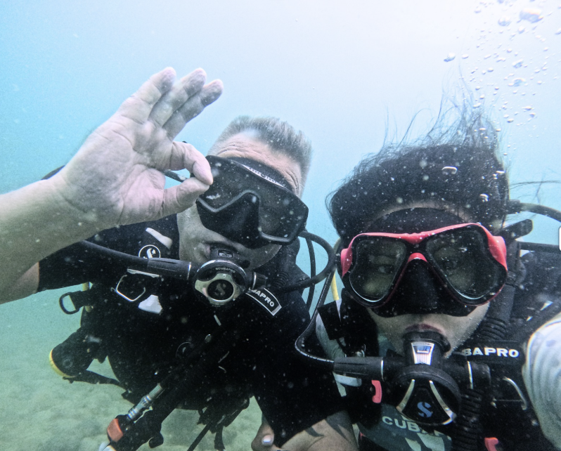 zwei taucher unter Wasser, weiblich und männlich schauen in die Kamera und machen das okay zeichen mit der hand